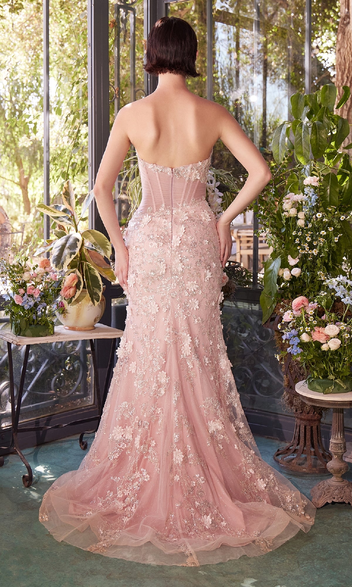 A woman in the Andrea & Leo A1396 strapless floral prom gown stands indoors facing away, surrounded by lush plants and large windows.